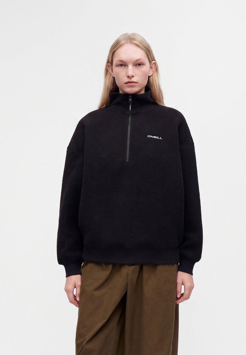 Jeune femme aux longs cheveux blonds porte une polaire noire zippée avec le logo O'Neill et un pantalon marron ample, debout devant un fond uni.