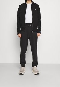 Chaqueta negra con cremallera, cuello alto y bolsillos, combinada con pantalones jogger negros con cordón ajustable. Zapatillas deportivas grises y beige.