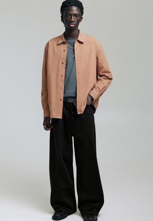 Nuori mies, jolla on tumma iho ja punotut hiukset, pukeutuneena beigeihin ruudullisiin paitaan harmaan t-paidan päällä, leveisiin mustiin housuihin ja kerroksittaisiin hopeisiin kaulakoruihin.