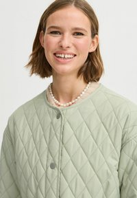 Chaqueta acolchada de color verde claro con cierres de botón y una tela suave y texturizada. Llevada sobre una camisa ligera y texturizada con un collar de perlas.