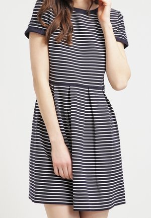 Femme portant une robe à manches courtes rayée horizontale bleu marine et blanc avec une jupe plissée, debout devant un fond uni.