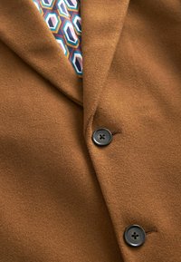 Manteau en laine marron avec deux boutons noirs attachés, dévoilant partiellement une chemise aux motifs géométriques colorés en dessous.