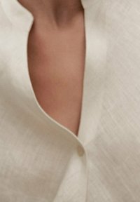 Linned blouse med et subtilt tekstureret mønster, en blød cremefarve og en rund krave med en enkelt knaplukning.