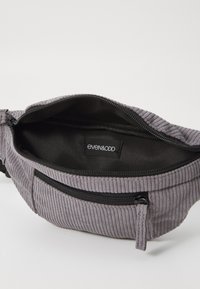 Harmaa corduroy vyölaukku mustalla sisäosalla, jossa on päävetoketjullinen osasto ja edessä vetoketjullinen tasku. Brändilogo näkyvissä.