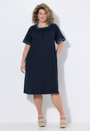 Kobieta o kręconych włosach, ubrana w granatową sukienkę do kolan z wzorzystą lamówką i beżowe sandały na platformie, stojąca na białym tle.