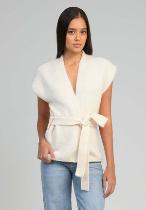 Femme portant un gilet sans manches blanc et pelucheux avec des épaules larges et une ceinture nouée, associée à un jean bleu clair sur un fond uni.