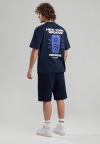 Navy T-shirt with graphic design of speakers and text "NEW YORK GROOVE RECORDS" in white and blue, paired with navy shorts and white sneakers.