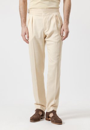 Hombre con pantalones plisados beige con hebilla lateral y sandalias de cuero trenzado marrón oscuro contra fondo blanco.