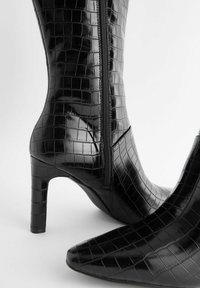 Črne škornje do kolen z krokodiljim vzorcem, z nazobčano konico in zadrgo. Sijajna površina in peto v stiletto stilu poudarjata eleganten dizajn.