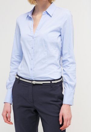 Mujer con una camisa azul claro abotonada metida en unos pantalones azul marino oscuro con un cinturón fino beige, de pie sobre un fondo blanco.