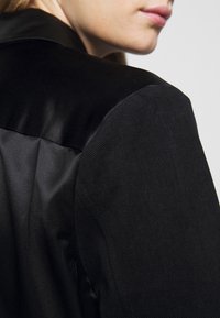 Svart blazer med en blandning av matta och glänsande ytor, strukturerade axlar och en slät satin-krage. Skräddarsydd passform med rena linjer.