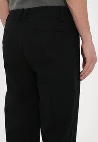 Homme portant un pantalon noir avec des passants de ceinture et une poche arrière, debout sur un fond clair uni, main reposant sur le côté.