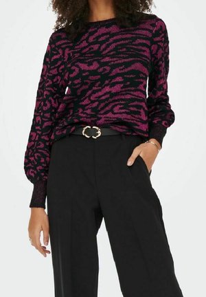 Femme portant un pull violet et noir à imprimé animal, rentré dans un pantalon noir taille haute avec une ceinture noire ornée d'une boucle dorée.