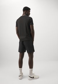 Camiseta atlética negra de manga corta y cuello redondo, combinada con pantalones cortos negros. Lleva calcetines blancos y zapatillas de deporte blancas, diseño minimalista.
