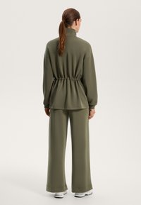 Loungewear set v olivno zeleni barvi vključuje top z visokim ovratnikom in pasom na vrvico ter široke hlače, izdelane iz mehkega, gladkega materiala z minimalnimi šivi.