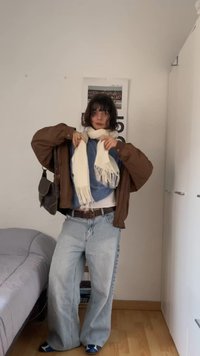 Νεαρή γυναίκα σε εσωτερικό χώρο, προσαρμόζει ένα λευκό μαντήλι, φορά καφέ σακάκι, μπλε μπλούζα, φαρδιά ανοιχτό μπλε τζιν και κρατά τσάντα ώμου δίπλα σε κρεβάτι και ντουλάπα.