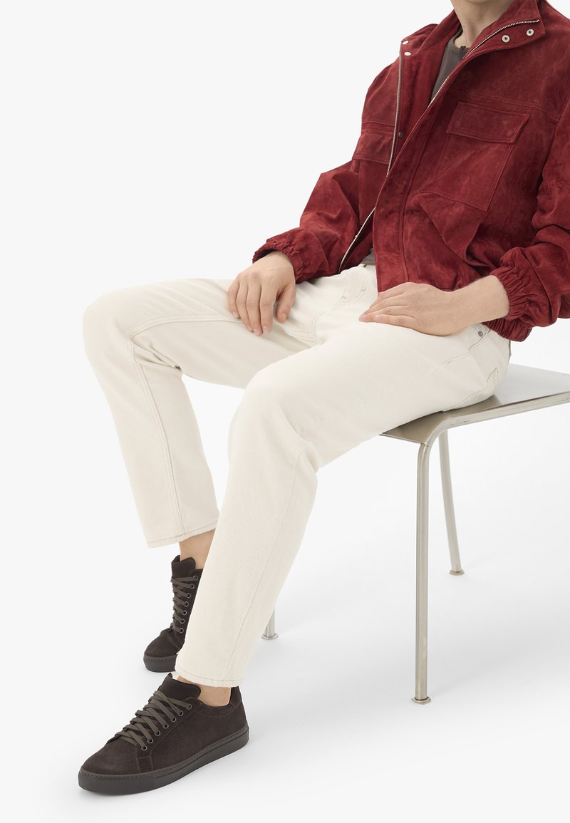 Personne assise sur une chaise en métal portant une veste rouge, un pantalon beige clair et des baskets noires avec des lacets sur un fond blanc.