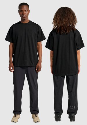 Schwarzes, übergroßes T-Shirt mit rundem Halsausschnitt und kurzen Ärmeln. Getragen mit dunklen Hosen, die Reißverschlüsse und ein graues numerisches Detail am Bein aufweisen.