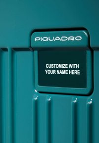 Tegelkleurige harde koffer met geribbelde patronen, voorzien van een aanpasbare naamplaat met het "Piquadro"-logo en de tekst "CUSTOMIZE WITH YOUR NAME HERE".