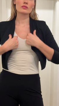 Donna che aggiusta il suo blazer nero sopra una maglia bianca, in piedi contro una parete chiara e liscia.