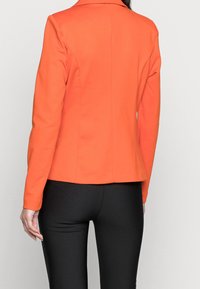 Pomarańczowa dopasowana marynarka z długimi rękawami, gładką fakturą i czystym szwem z tyłu, zestawiona z czarnymi dopasowanymi spodniami.