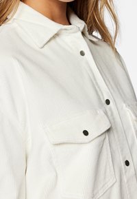 Biała koszula sztruksowa z kołnierzykiem, dwiema przednimi kieszeniami, zapięciem na zatrzask oraz subtelną pionową teksturą prążków.