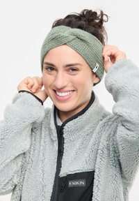 Grünes gestricktes Kopfband mit einem verdrehten Design; weiche Textur. Getragen mit einer grauen, flauschigen Jacke mit Reißverschluss, die ein schwarzes Panel und ein Logo aufweist.