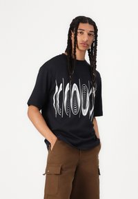 Svart bomullströja med ett grafiskt mönster av vita vågor, korta ärmar och avslappnad passform, i kombination med bruna cargoshorts.