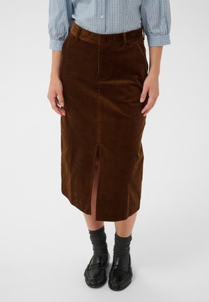 Personne portant une jupe midi en velours côtelé marron avec une fente devant, des chaussettes grises, des mocassins noirs et une chemise à carreaux bleue à manches longues, debout sur un fond blanc.