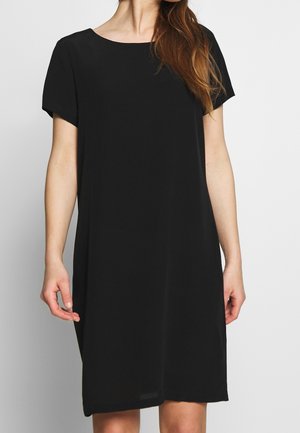 Femme portant une robe noire unie à manches courtes, longueur genou, avec un col rond, debout devant un fond uni.