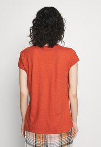 Kvinde med krøllet mørkt hår iført en kortærmet orange bluse og et rute-mønstret nederdel, vendt væk mod en ensfarvet lys baggrund.