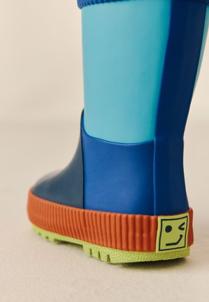 Bota de lluvia azul y turquesa con suela acanalada naranja y un logo cuadrado amarillo con una cara guiñando el ojo en el talón.