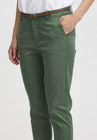 Chinos verts avec une texture lisse, coupe slim et poches latérales. Dotés d'une ceinture marron avec une boucle en métal. Associés à une chemise blanche.