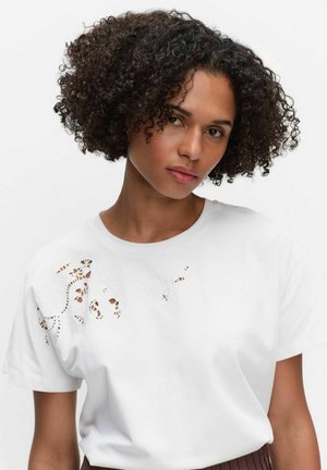 Kvinde med kort krøllet hår iført en hvid T-shirt med blomsterudskæringsmønstre på skulderen og øvre bryst.