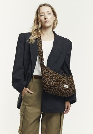 Brown leopard print shoulder bag with a wide strap and a logo patch, made from textured fabric, worn with a dark oversized blazer and khaki pants.