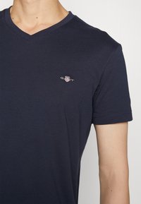 Mężczyzna w granatowym T-shircie z dekoltem w serek i małym logo GANT na lewej piersi.