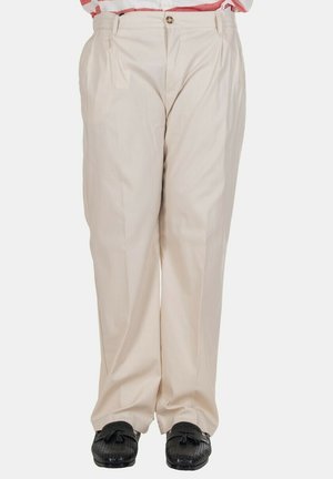 Pantaloni beige plissettati indossati con una camicia a righe parzialmente visibile e mocassini neri intrecciati con nappine su sfondo bianco.