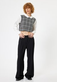 Sort-hvidternet cropped vest over en hvid langærmet skjorte, parret med sorte vide bukser og sko. Subtile teksturdetaljer til stede.