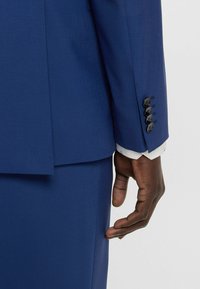 Marineblauw colbert met een soepele textuur, voorzien van zwarte knopen op de manchetten. Een witte shirthals is zichtbaar onder de mouwen van het colbert.