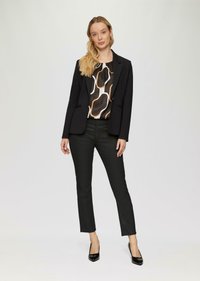 Schwarze Blazer über einer gemusterten Bluse in Braun, Weiß und Orange, kombiniert mit schwarzen Slim-Hosen und schwarzen spitzen Schuhen.