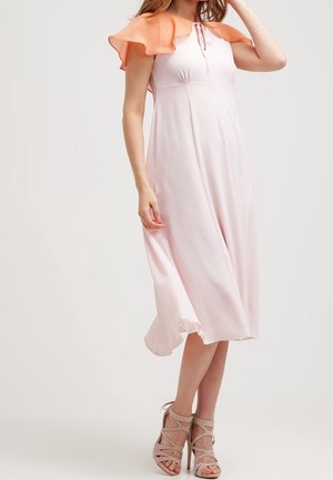 Femme portant une robe midi rose clair avec un capeline corail à volants, assortie à des sandales à talons hauts beiges à brides.