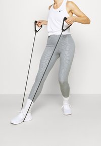 Leggings gris clair à motif texturé, associées à un débardeur blanc. Bandes de résistance avec poignées noires tenues en main, portées avec des baskets blanches.