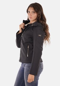 Giacca nera con cappuccio, chiusura con zip sul davanti, tasche laterali e un emblema dorato sul braccio. Realizzata in un materiale liscio e resistente alle intemperie.