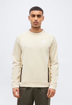 Uomo che indossa una felpa beige North Face con tasche laterali con cerniera e pantaloni scuri, in piedi davanti a uno sfondo semplice.