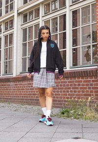 Black bomber jacket with a blue emblem, white t-shirt, plaid skirt, turquoise sneakers with red accents, and white socks. Urban setting.