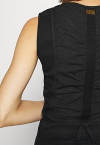 Czarny top bez rękawów z marszczonym wzorem z tyłu, posiadający centralny szew i kontrastowe czarne ramiączka. Materiał wydaje się gładki i lekko błyszczący.