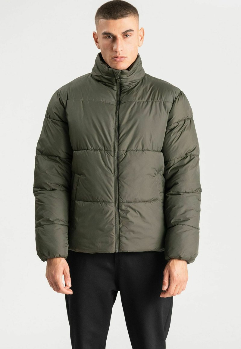 Homme aux cheveux courts portant une doudoune vert foncé et un pantalon noir, debout devant un fond blanc uni.