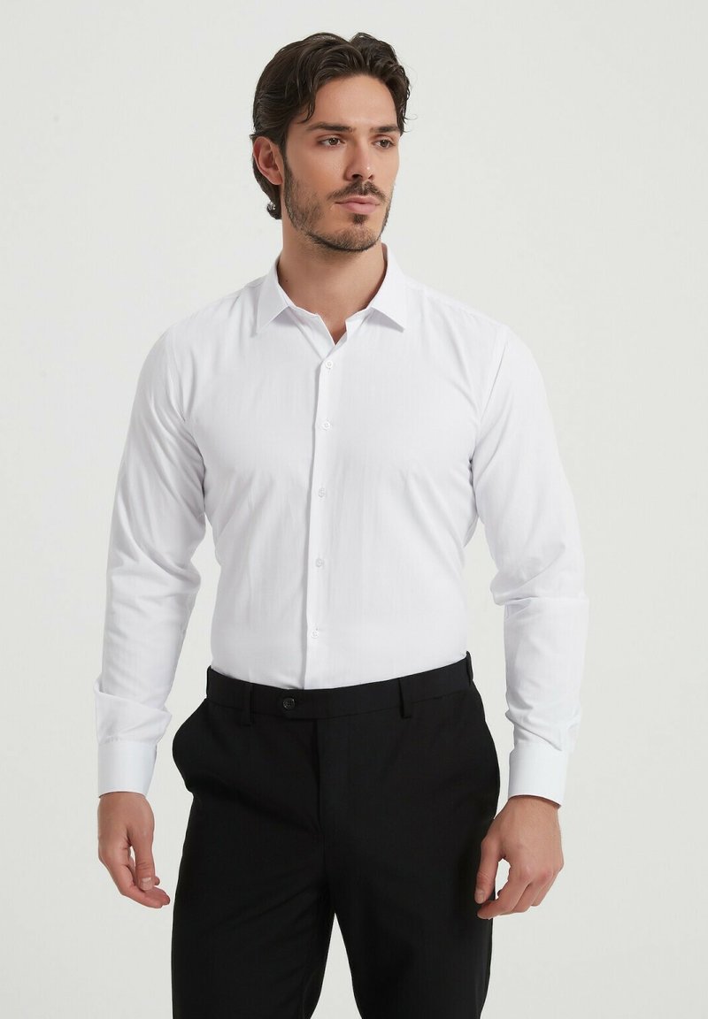 Chemise blanche à manches longues avec un col pointu, fermeture à boutons et texture lisse, associée à un pantalon noir.