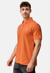 Oranje polo-shirt met een kraaghals, korte mouwen en een subtiel logo op de borst. Draag het met donkergrijze jeans.