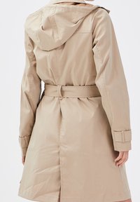 Beige trench coat with hood, belted waist, and adjustable wrist straps worn by a person shown from the back against a plain background.